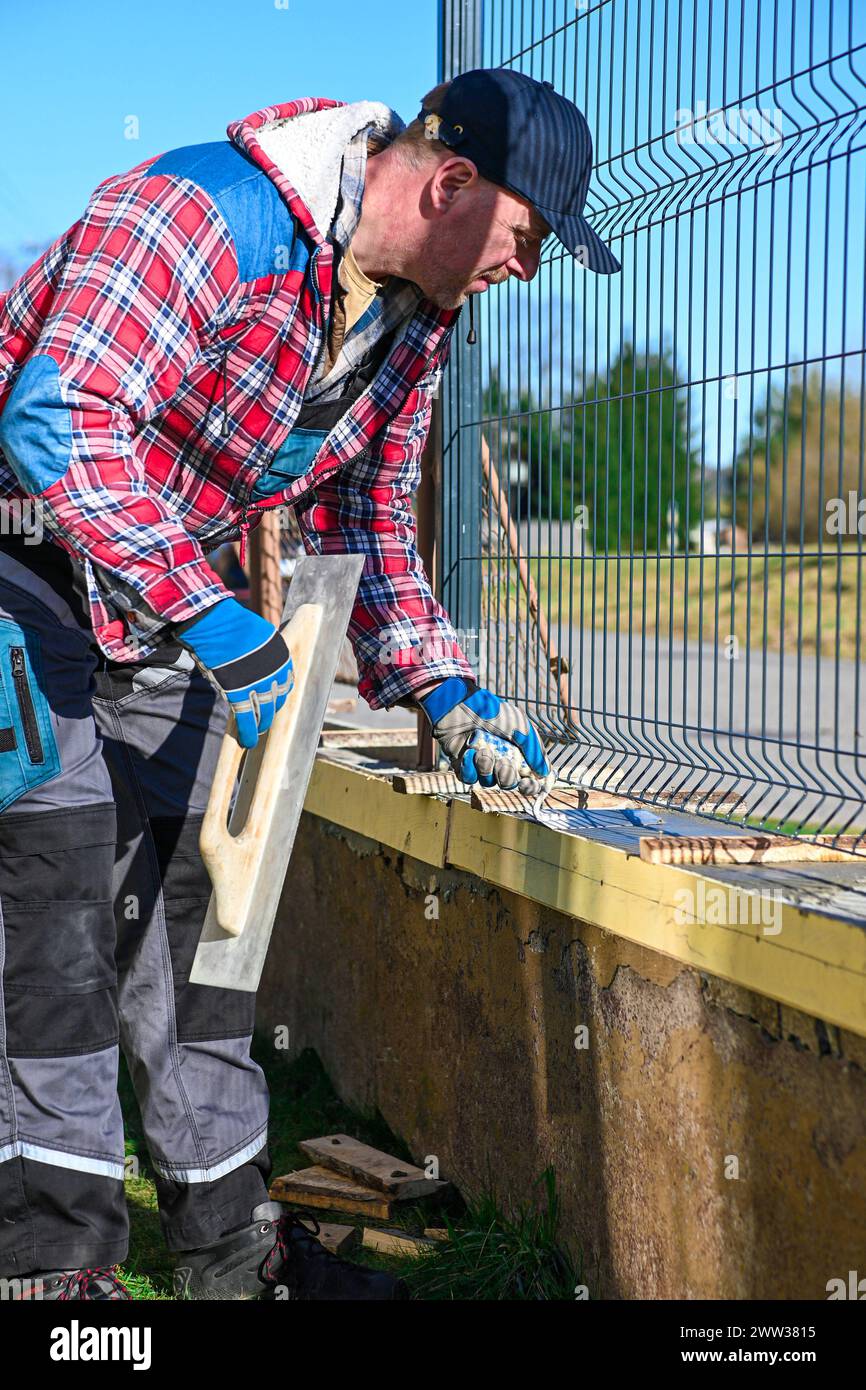 Ein Mann in Overall und Handschuhen repariert den Zaun vor dem Familienhaus. Nahansicht und unscharfer Hintergrund. Stockfoto