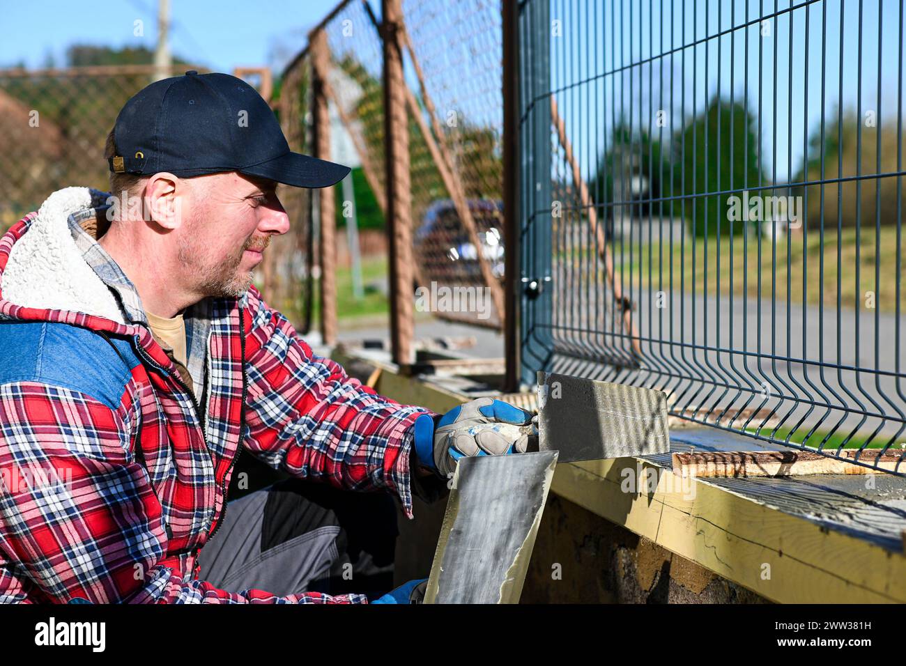 Ein Mann in Overall und Handschuhen repariert den Zaun vor dem Familienhaus. Nahansicht und unscharfer Hintergrund. Stockfoto