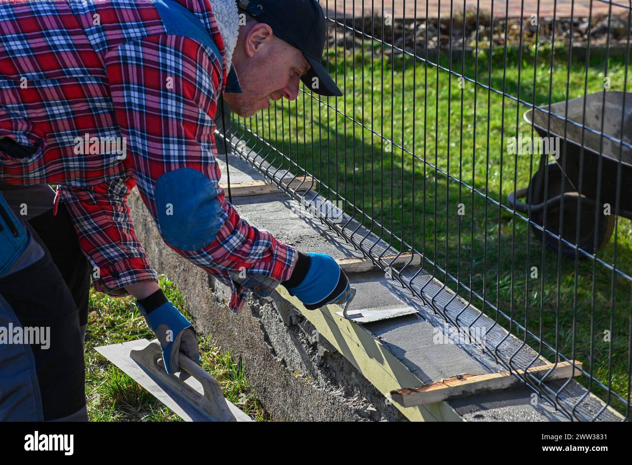 Ein Mann in Overall und Handschuhen repariert den Zaun vor dem Familienhaus. Er benutzt ein Flugzeug, eine Kelle und eine Schubkarre. Stockfoto
