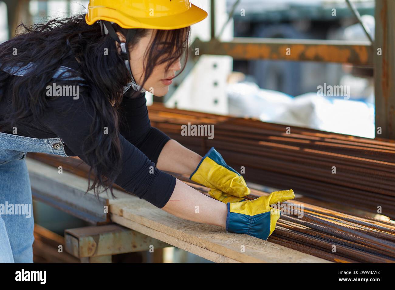 Entschlossene hispanische Frau auf der Baustelle, die Sicherheitsausrüstung trägt, die Stahlstäbe mit Schutzhandschuhen umgeht Stockfoto