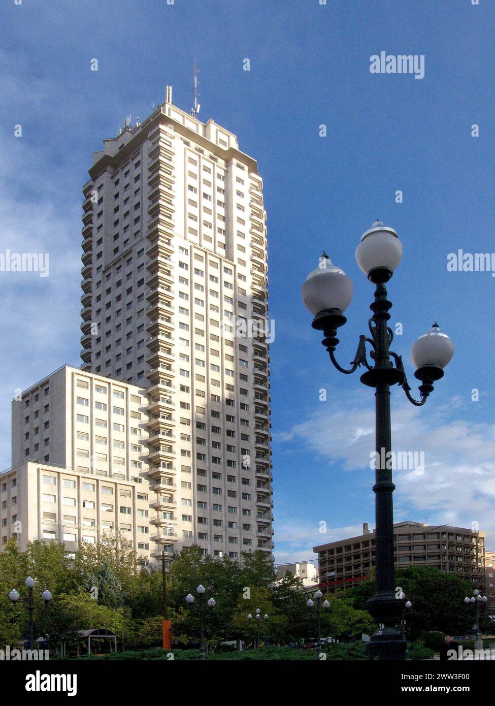 Ein Wolkenkratzer erhebt sich in den blauen Himmel, neben einer klassischen Straßenlaterne und umgeben von Grün Madrid Spanien Stockfoto