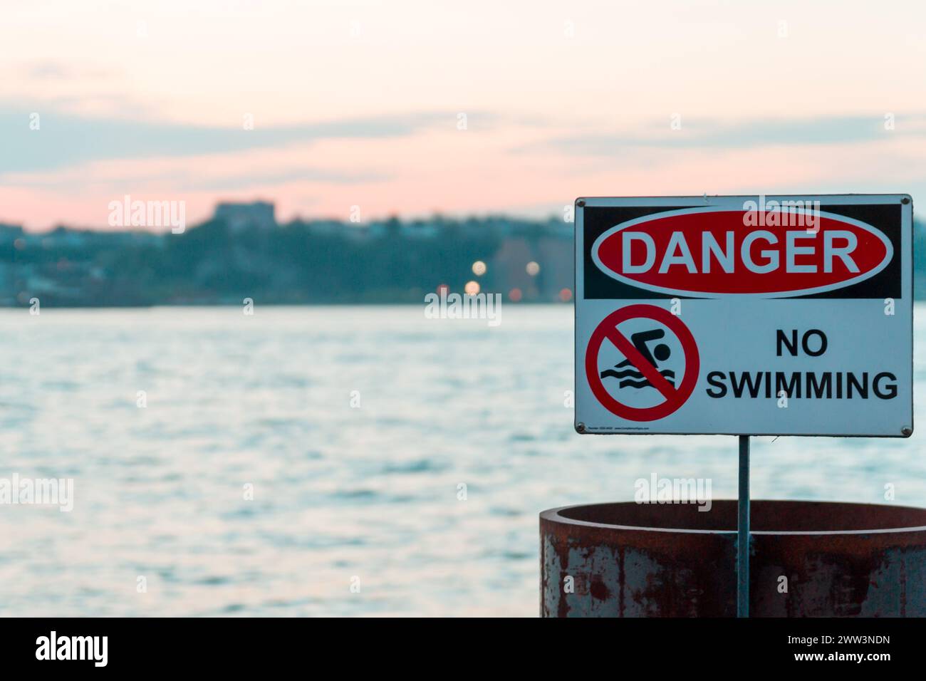 Warnschild „Gefahr kein Schwimmen“ am Meer mit einer rostigen Barriere und Hintergrund des Abendhimmels Stockfoto