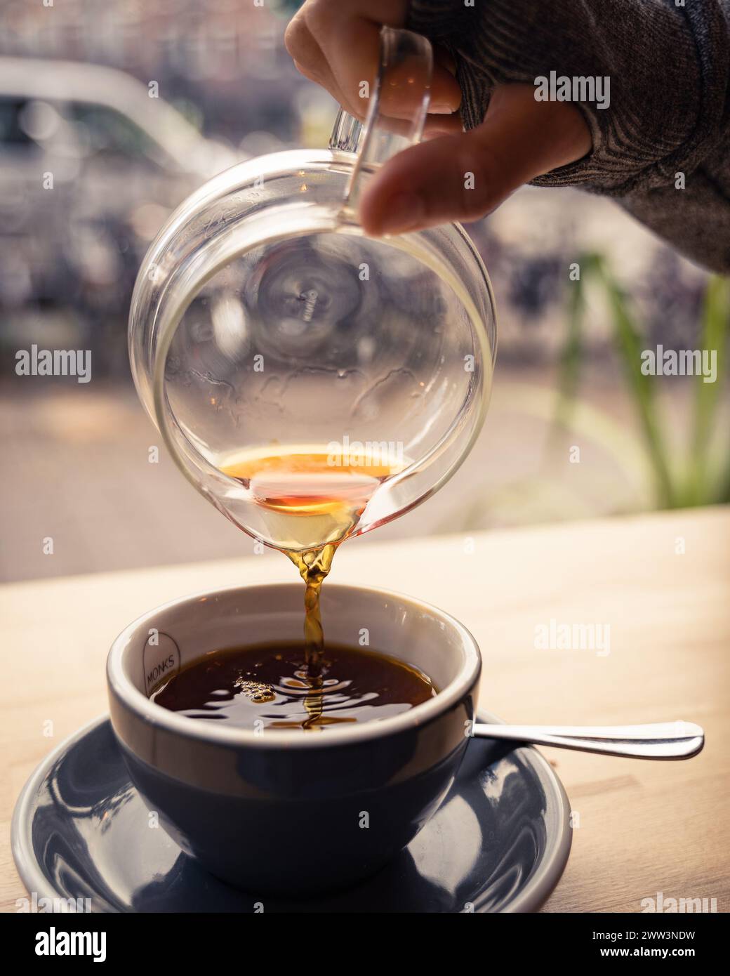 Nahaufnahme einer Hand, die an einem Holztisch in einem gemütlichen Café dampfenden Kaffee in eine Tasse gießt Stockfoto