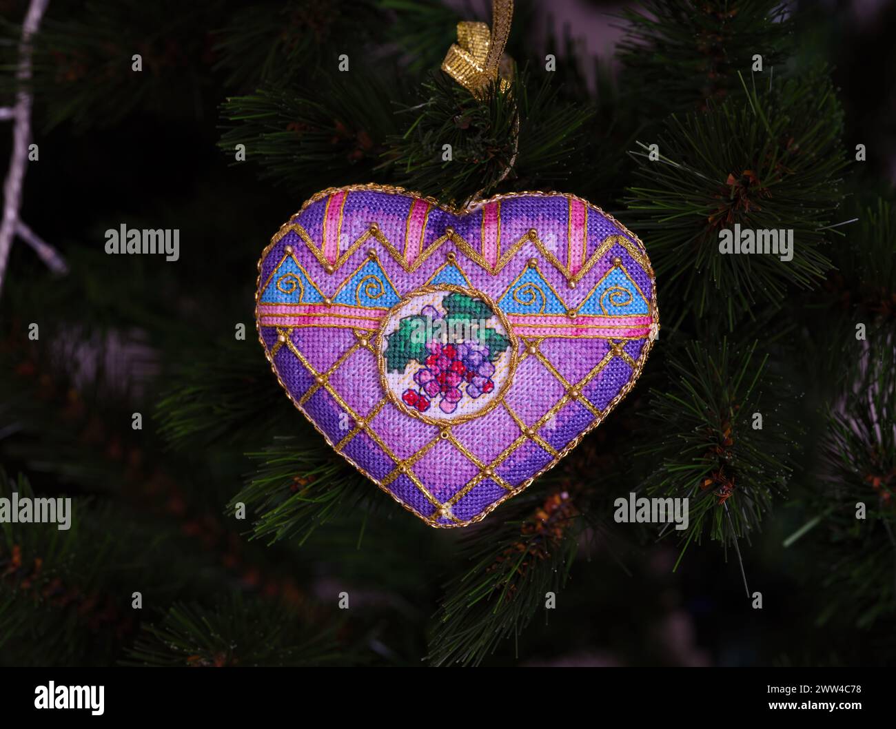 Ein kreuzgenähtes zeitloses Weihnachtsschmuck auf einem Weihnachtsbaum. Dieses lila Weihnachtsschmuck in Herzform mit aufgestickten Trauben und m Stockfoto