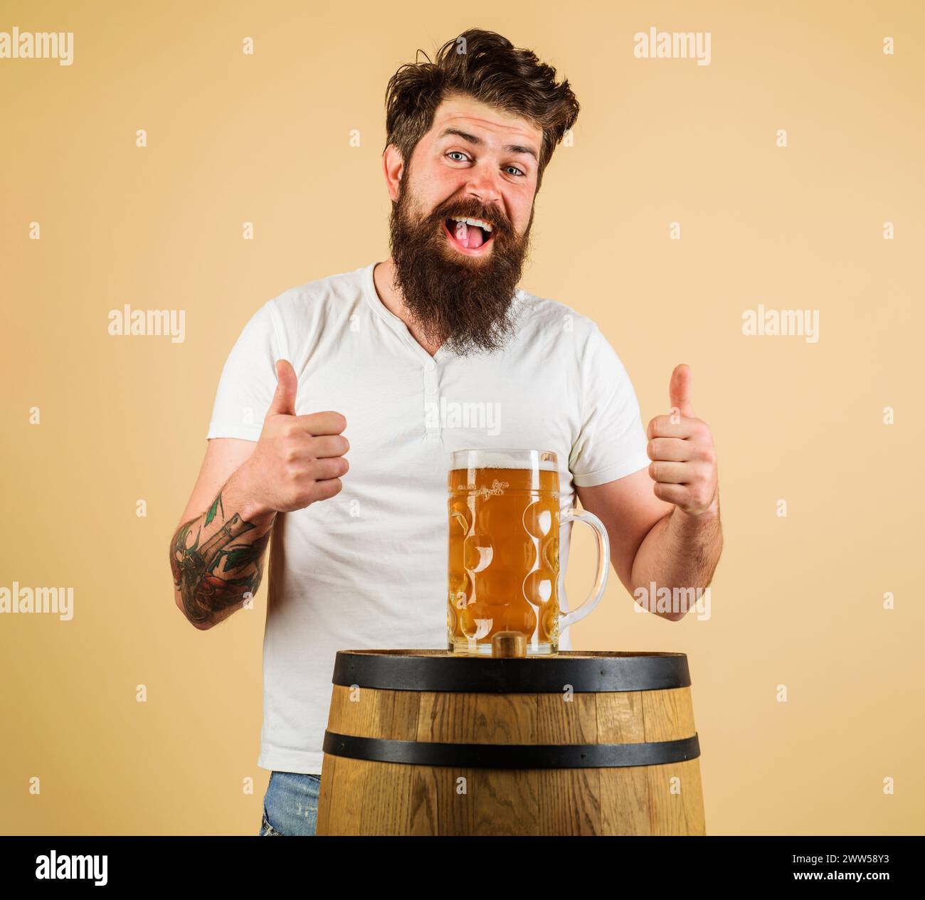 Bärtiger Mann mit einem Becher Bier auf Holzfässern, der Daumen nach oben zeigt. Glücklicher Brauer mit einem Glas Lagerbier. Ausrüstung für die Zubereitung von Bier. Bierkneipe Stockfoto