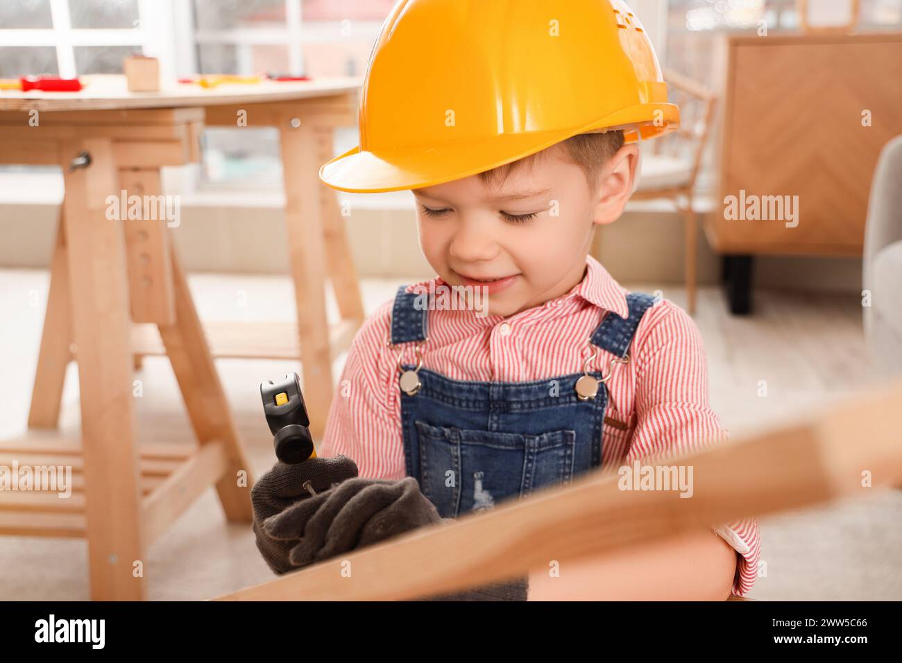 Niedlicher kleiner Baumeister, der Nagel in Holzplanke im Zimmer hämmert, Nahaufnahme Stockfoto