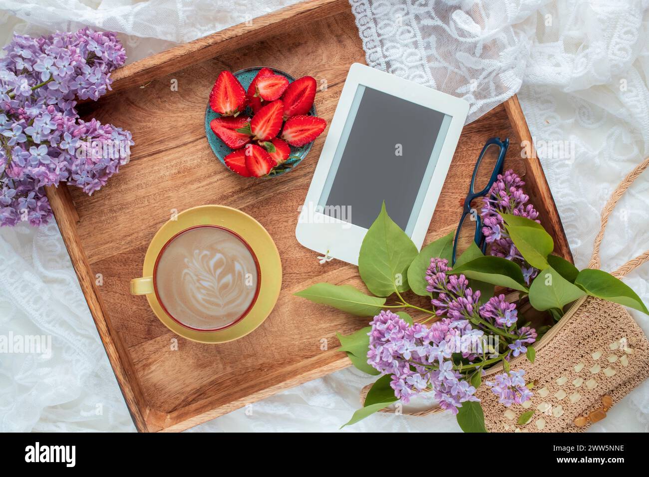 Blick von oben auf Kaffee im Bett mit leckeren Erdbeeren, E-Reader und lilafarbenen Zweigen auf Holztablett. Frühling, Sommer Stillleben Komposition. Stockfoto