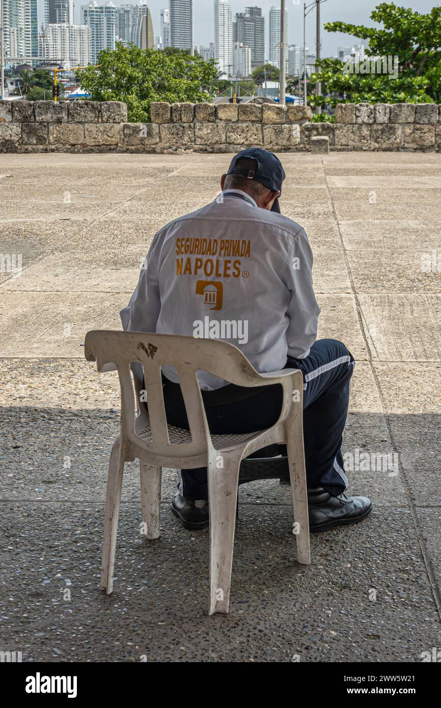 Cartagena, Kolumbien - 25. Juli 2023: Privater Sicherheitsbeamter sitzt auf einem Plastikstuhl auf der Stadtmauer und Bastion von Baluarte de San Ignacio. Modernes hohes Gebäude Stockfoto