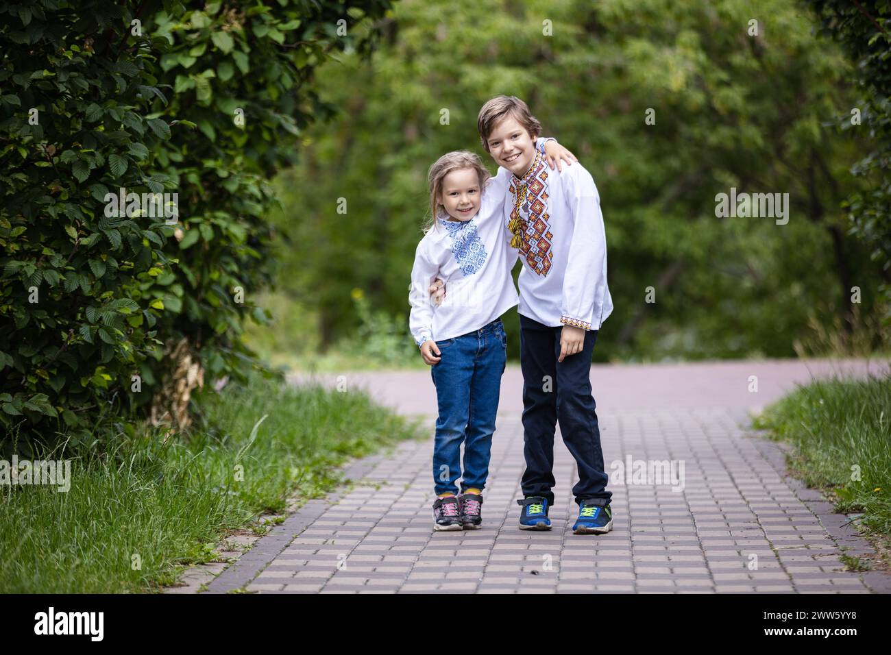 Kleine Kinder tragen traditionelle ukrainische bestickte Hemden, die sich umarmen, während sie in die Kamera schauen. Bruder und Schwester posieren draußen im Park Stockfoto