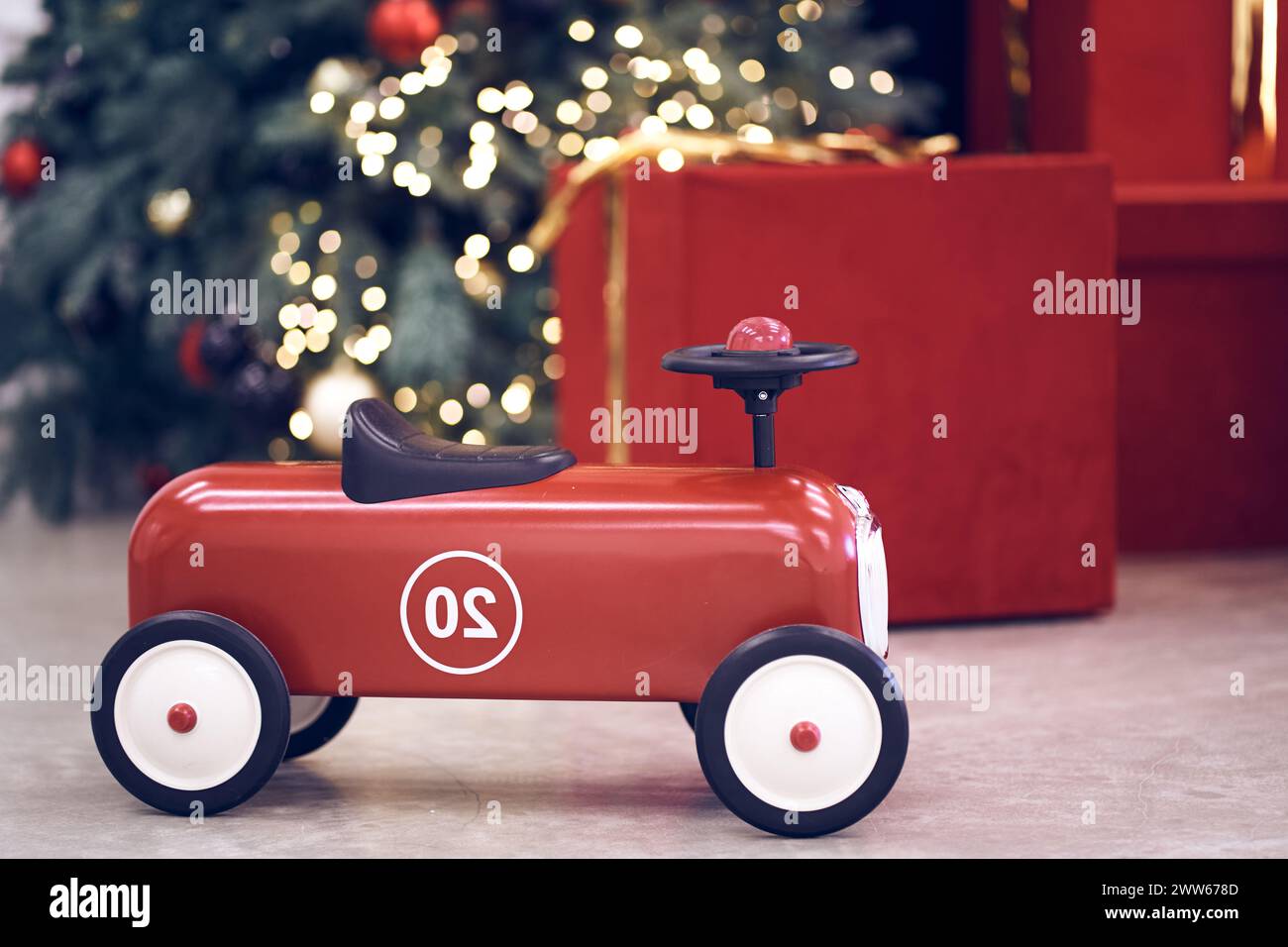 Blick auf das rote Retro-Auto für Kinder auf dem Hintergrund von Geschenken in roten Schachteln mit goldenen Bögen, Weihnachtsbaumgirlande, Lichterketten, roten und blauen Kugeln. Hintergrund, Kopierraum, Bokeh. Stockfoto
