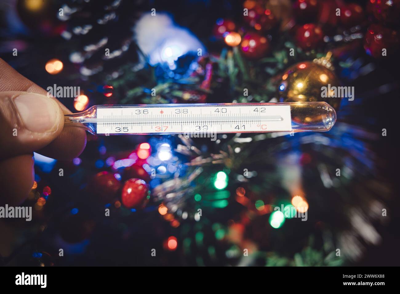 Eine Hand, die ein Thermometer auf dem Hintergrund verschwommener Lichter und weihnachtsbaumschmuck hält Stockfoto