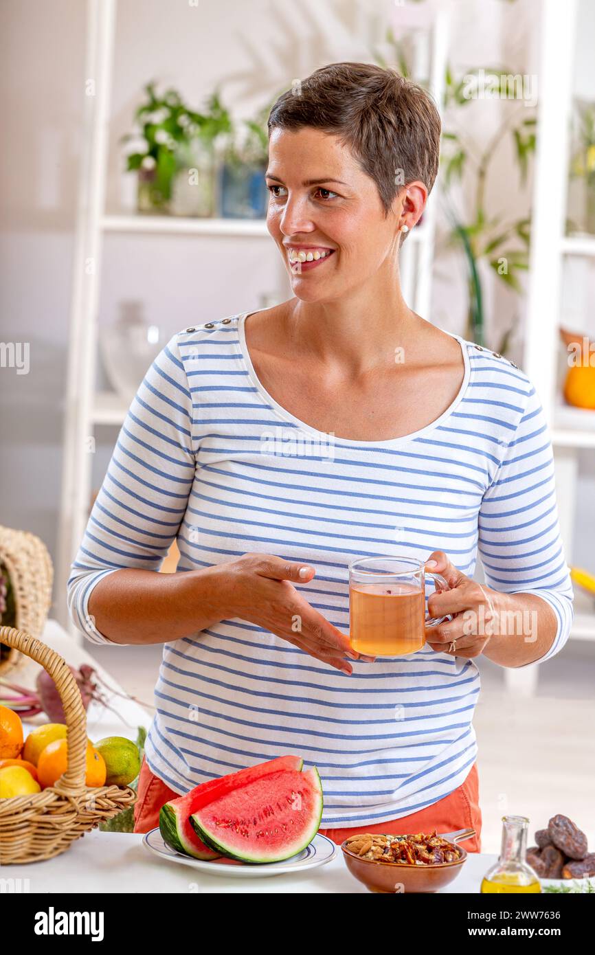 Frau, eine Tasse Tee in der Hand, umgeben von Getreide, frischen Früchten und getrockneten Früchten, Honig. Stockfoto