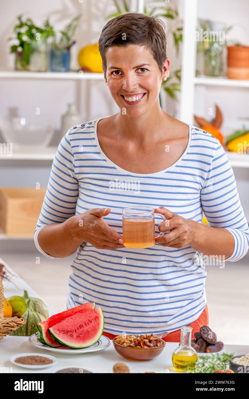 Frau, eine Tasse Tee in der Hand, umgeben von Getreide, frischen Früchten und getrockneten Früchten, Honig. Stockfoto