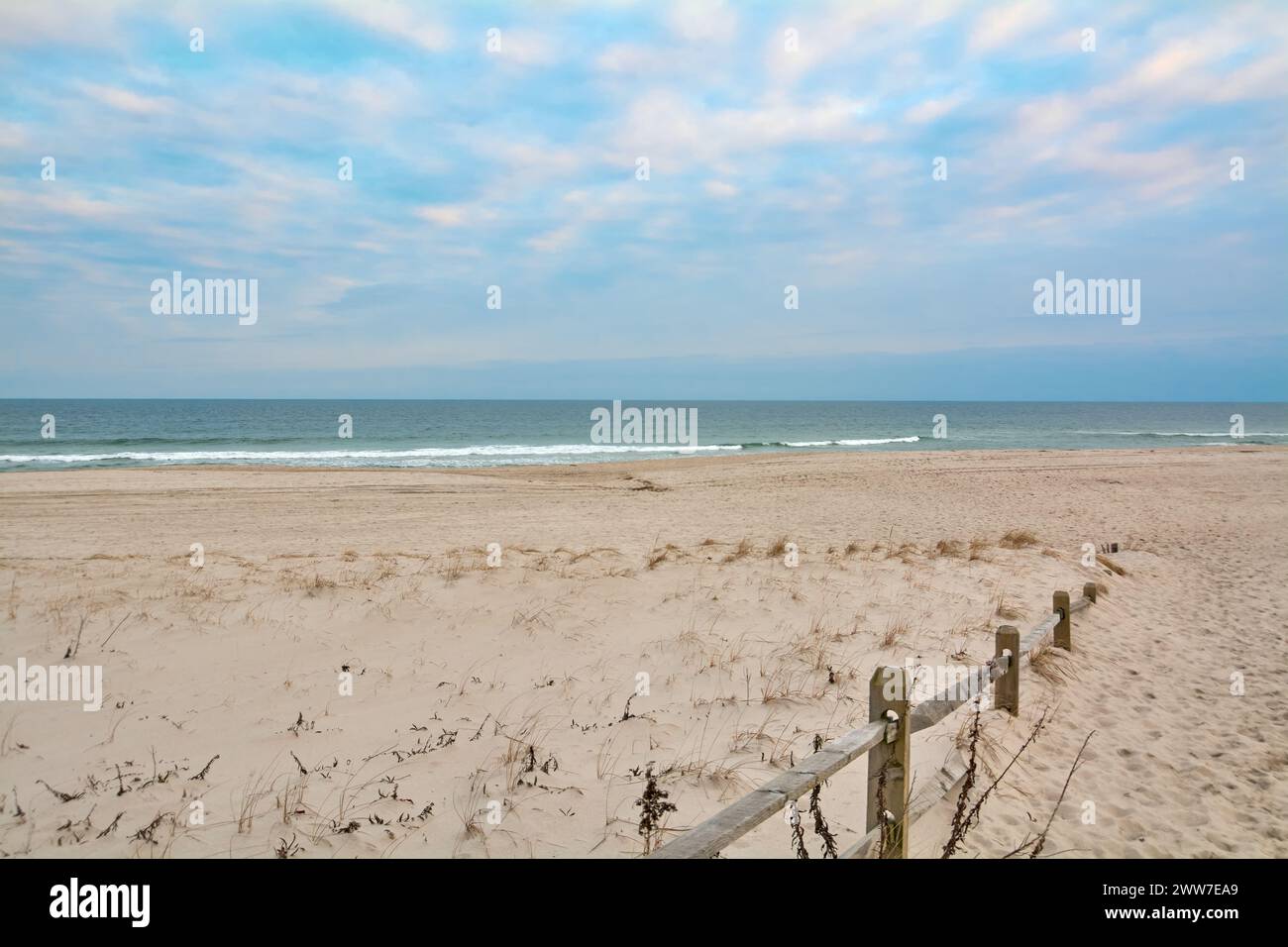 Ein sandiger Pfad führt uns durch die Küstenvegetation zu einem ruhigen Strand, mit einem Holzzaun, der eine rustikale Note verleiht. Stockfoto