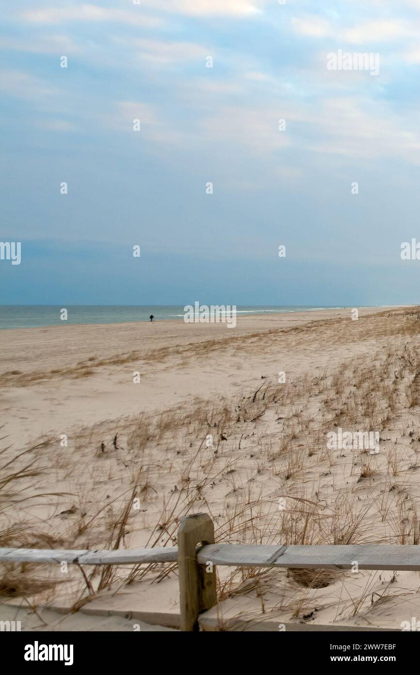 Das Bild zeigt einen sandigen Pfad, der zu einem ruhigen Strand führt, mit einem Holzzaun und einer Küstenvegetation, die einen rustikalen Charme verleiht. Stockfoto