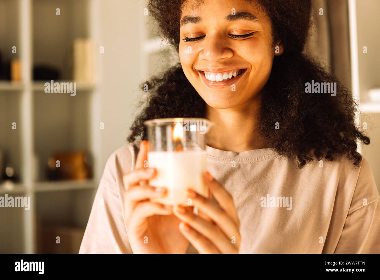 Nahaufnahme eines charmanten Teenagers, der eine Kerze in einem Glaskerzenleuchter hält und lacht. Ein hübsches afrikanisches Mädchen steht in einem stilvollen, hellen Ankleidezimmer Stockfoto