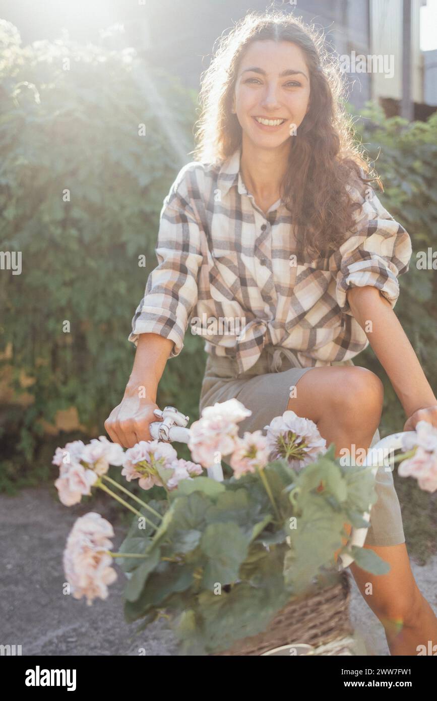 Eine junge attraktive Frau in lässiger Kleidung sitzt im Garten auf einem Fahrrad. Ein charmantes Mädchen mit lockigen Haaren lächelt draußen im hellen Sonnenli Stockfoto
