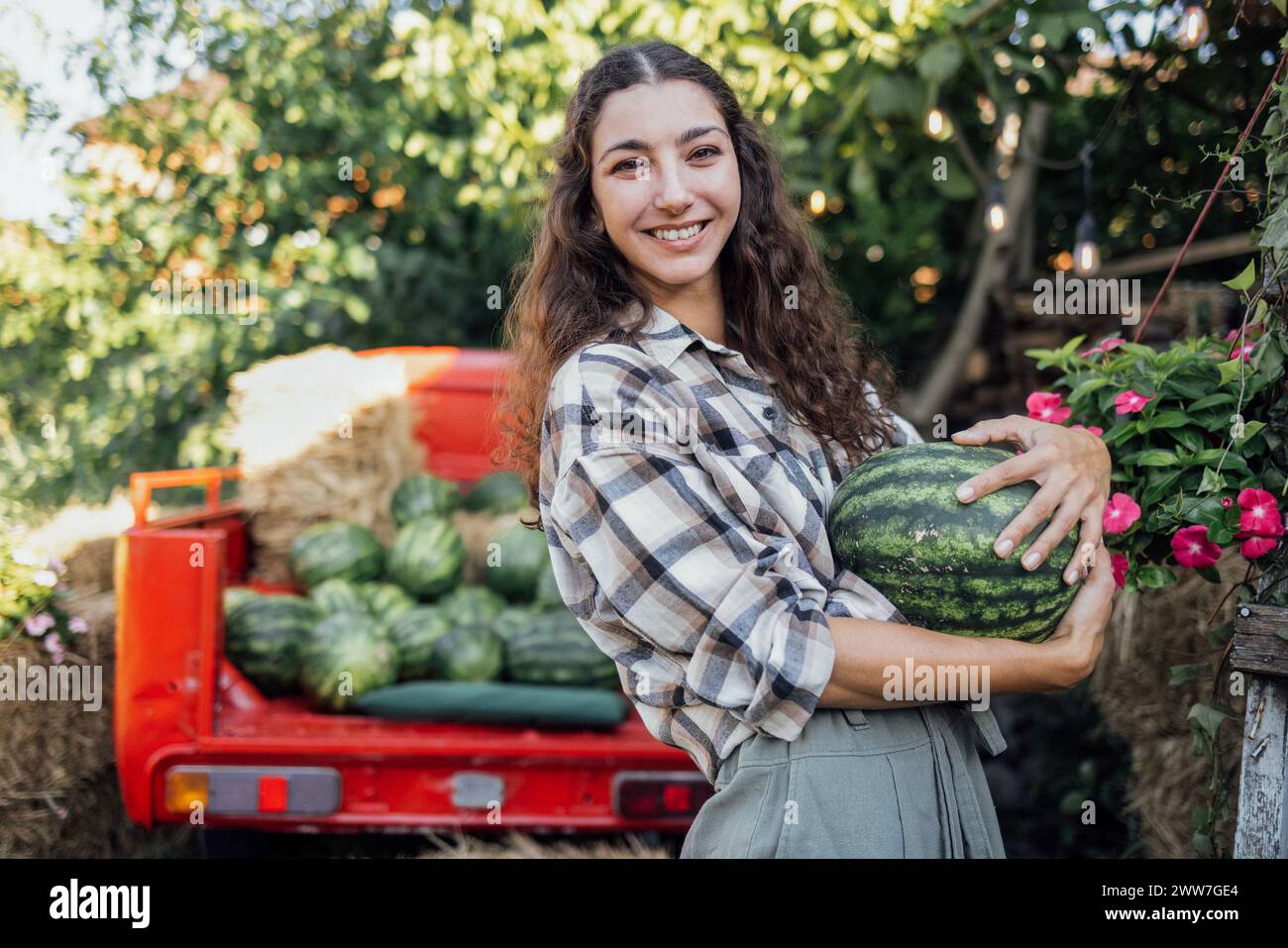 Charmante lächelnde Frau in lässigen Kleidern wirft Wassermelone draußen. Jungbauern mit lockigen Haaren sammeln frische, köstliche Beeren und Früchte. Ernte Stockfoto