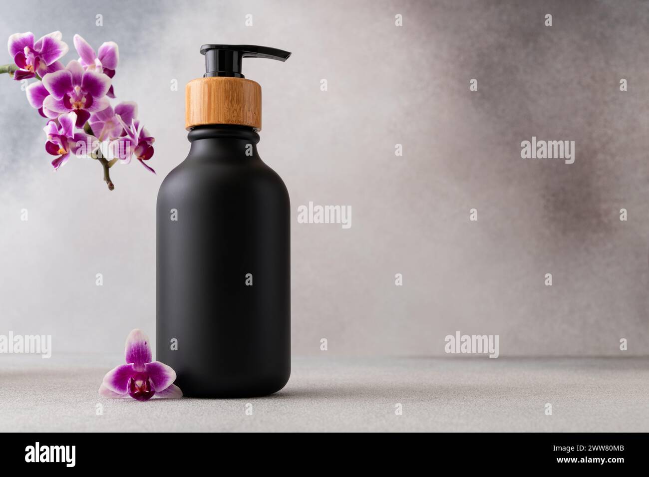 Schwarze, matte Flasche mit einem hölzernen Pumpdeckel und einer leuchtenden violetten Orchideenblüte für eine Spa-Atmosphäre. Die Szene erweckt ein Gefühl von Luxus und Ruhe Stockfoto