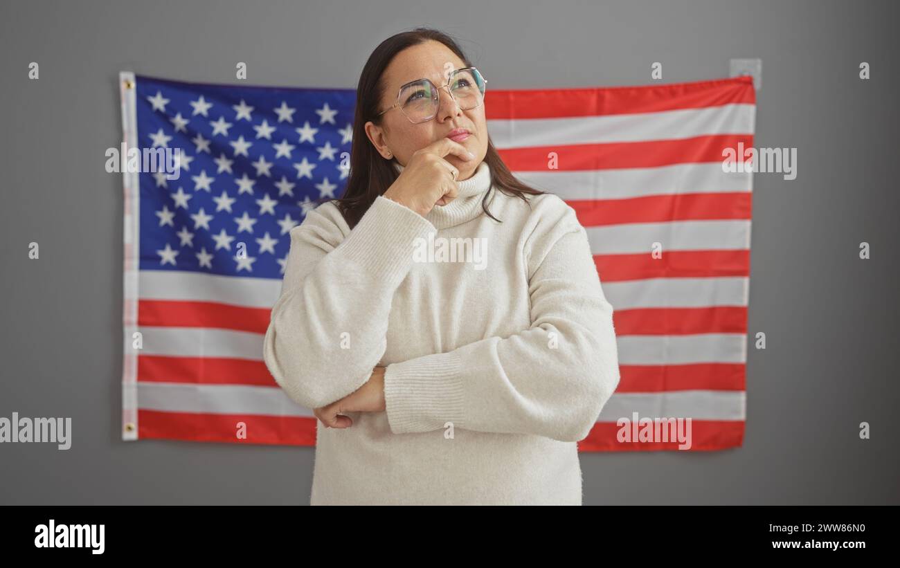 Nachdenkliche reife hispanische Frau im Amt mit usa-Flagge im Hintergrund Stockfoto