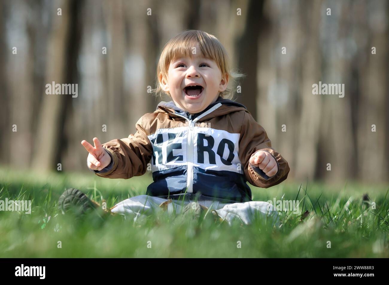 Glücklicher, süßer und lachender Junge, der Spaß hat, am Sonnenschein auf Gras zu sitzen. Freude und Glück Konzept. Liebe und familiäre Emotionen Stockfoto