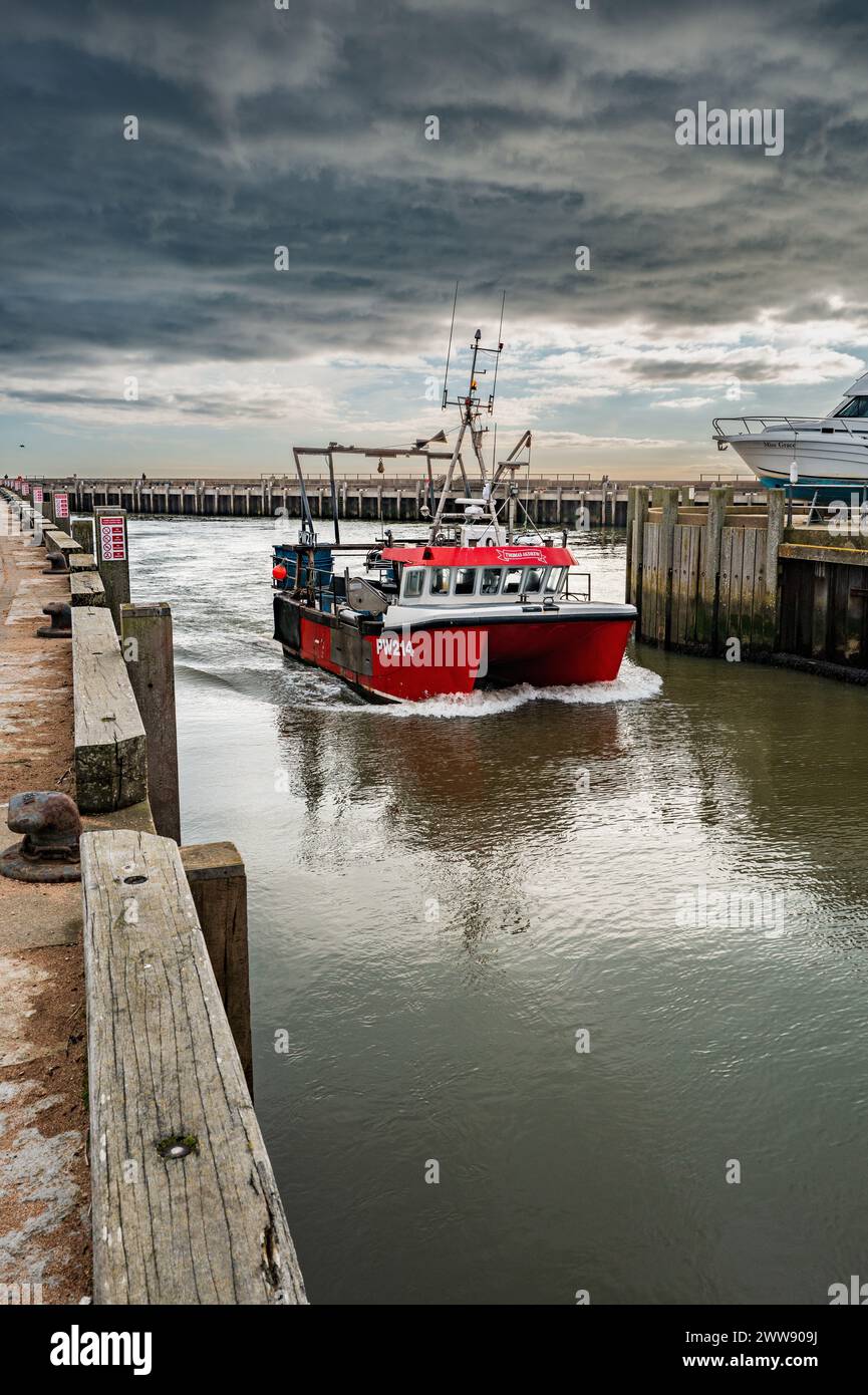 Das Fischerboot kehrt vor einer starken stürmischen Wolkenlandschaft zum Hafen von West Bay zurück. Am späten Nachmittag im Frühling. Kontrastfarben Rot und Grau. Stockfoto