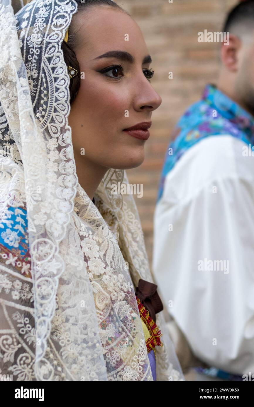 Valencianische Eleganz bei den Festlichkeiten von Gandia: Das weiße Spitzenkleid Einer Fallera, das mit traditionellen Mustern verziert ist, wird durch einen farbenfrohen Fan ergänzt Stockfoto