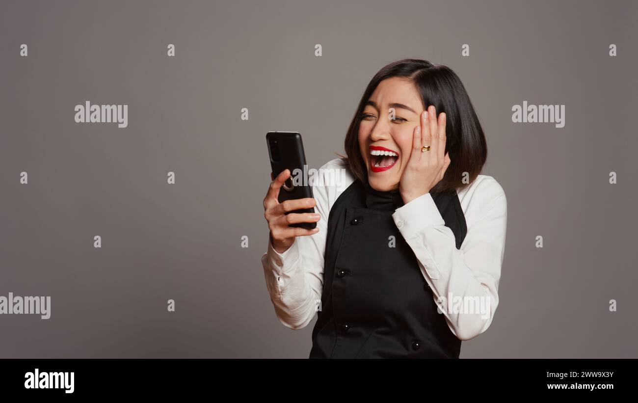 Der asiatische Kellner war zufrieden, nachdem er gute Nachrichten am Telefon gelesen hatte und viele Gäste reserviert hatten. Kellnerin mit Schürze, die mit dem Smartphone Tischreservierungen sieht, arbeitet gerne. Kamera B.. Stockfoto