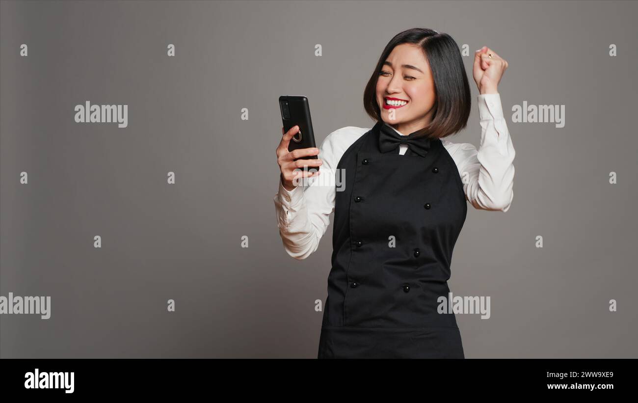 Der asiatische Kellner war zufrieden, nachdem er gute Nachrichten am Telefon gelesen hatte und viele Gäste reserviert hatten. Kellnerin mit Schürze, die mit dem Smartphone Tischreservierungen sieht, arbeitet gerne. Kamera A.. Stockfoto