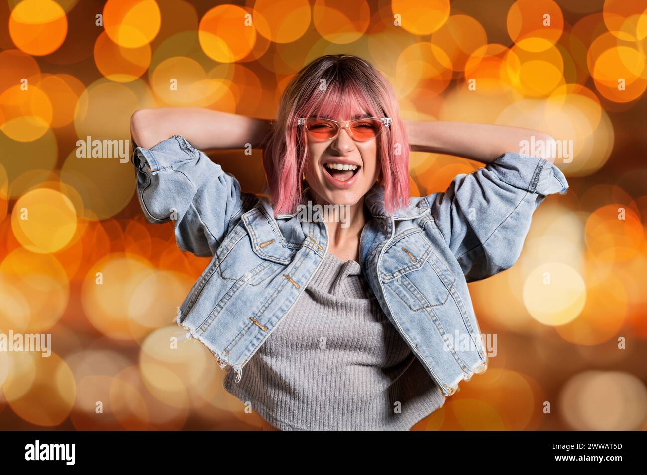Fröhliche, stilvolle Dame in Sonnenbrille und verkürzter Jeansjacke lacht mit Händen hinter dem Kopf auf orangefarbenem Bokeh-Hintergrund Stockfoto