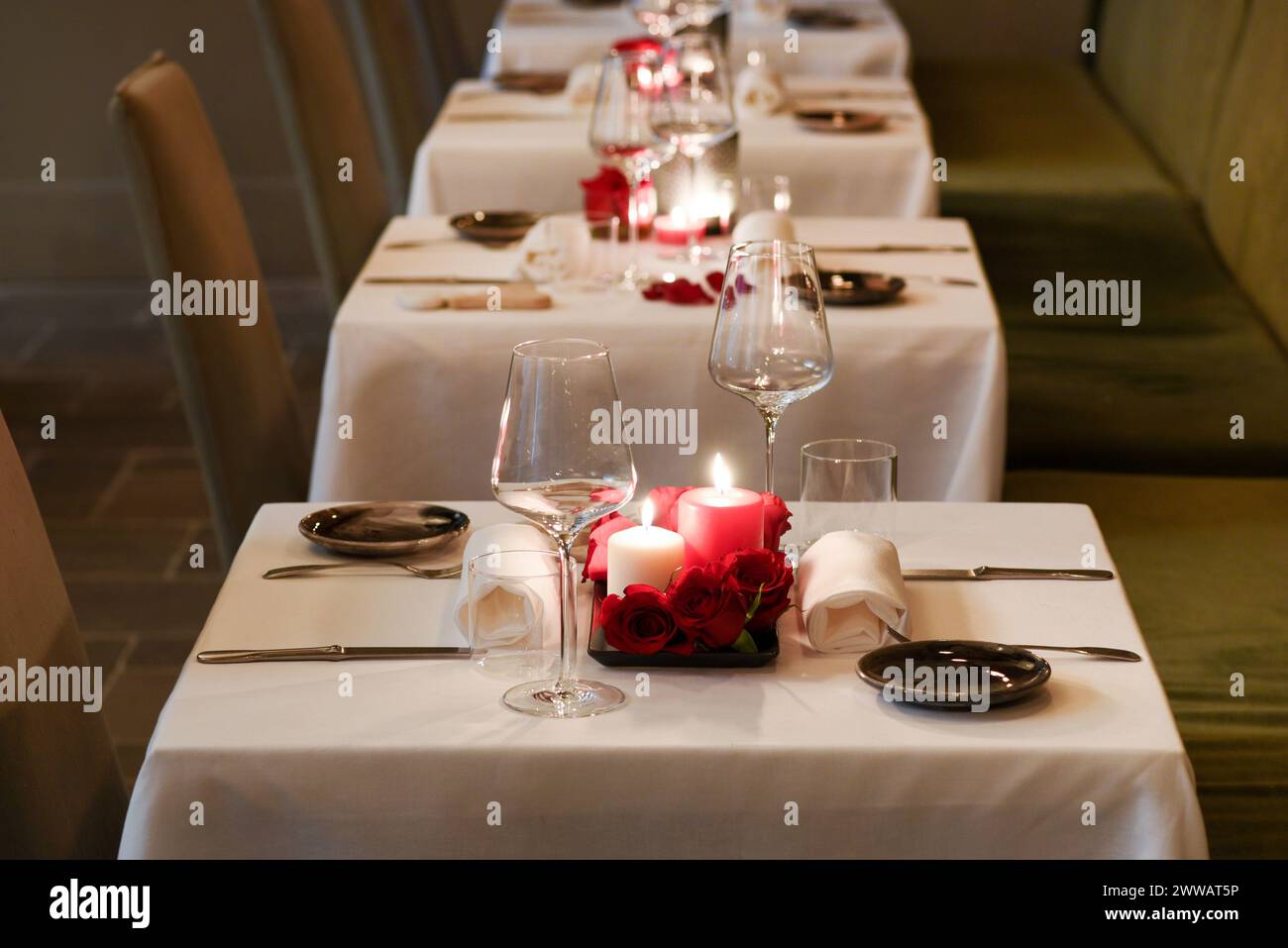 Reihe von Tischen, die mit brennenden Kerzen und leeren Weingläsern auf weißer Tischdecke im Luxusrestaurant dekoriert sind Stockfoto