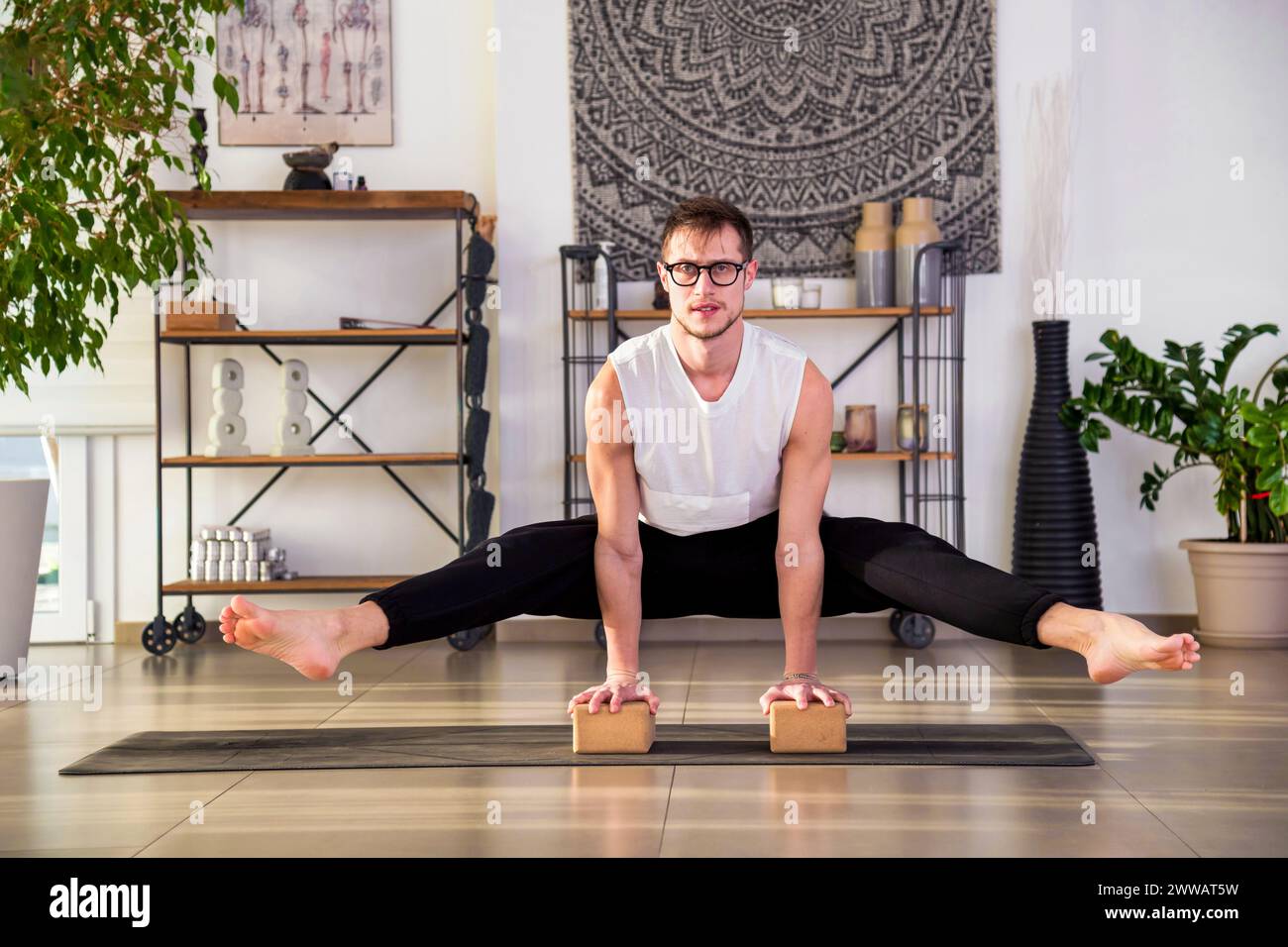 Der ganze Körper eines fokussierten Mannes in Sportbekleidung und Brille spreizt die Beine, während er während des Fitnesstrainings in einem modernen Studio mit pla-Topf Yoga-Blöcke hält Stockfoto