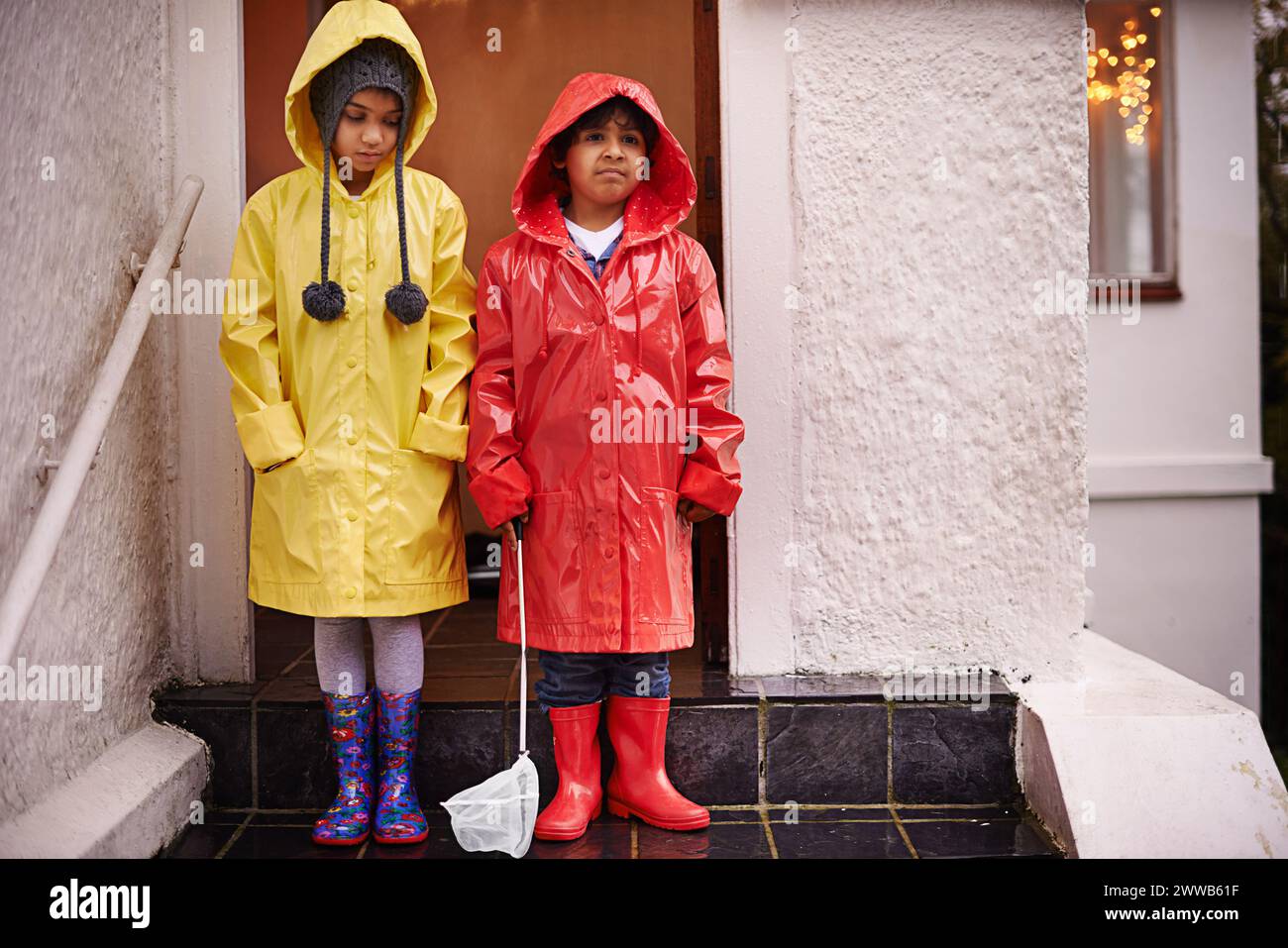 Kinder, Regenmantel und Porträt auf der Veranda für unglückliches, kaltes Wetter und das Aufwachsen für Jugendliche und Unschuldige. Kinder oder Kinder mit Stiefeln und verärgert mit Stockfoto