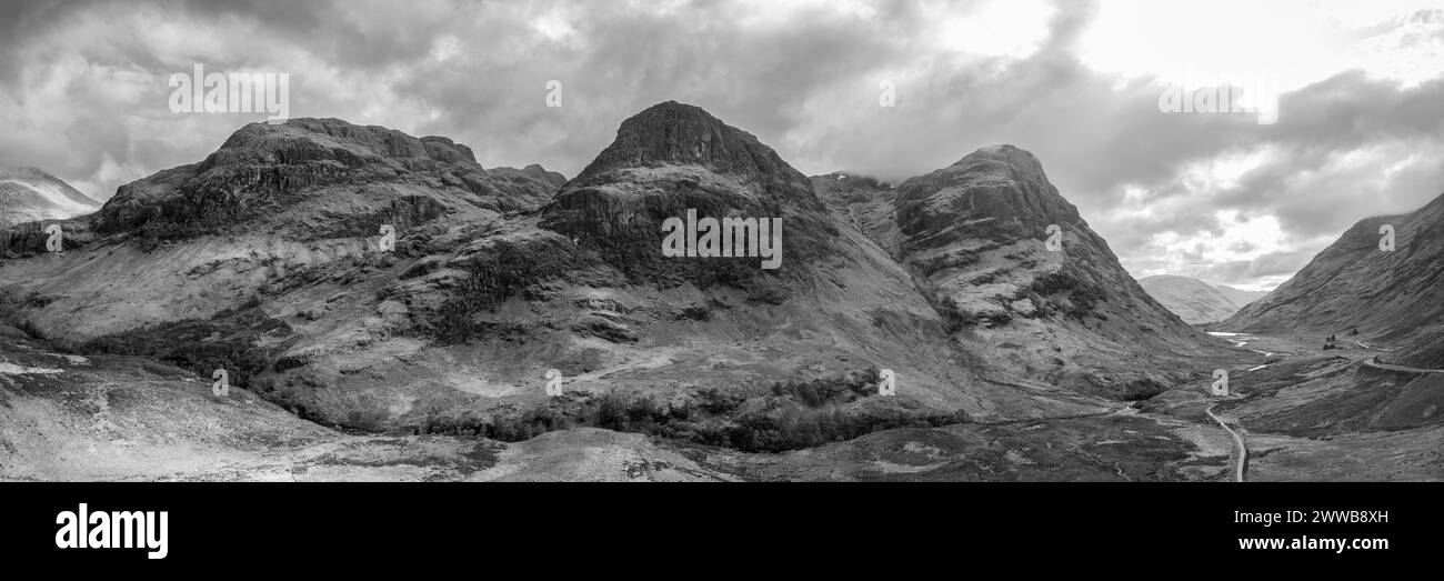 Dieses fantastische Panorama zeigt die majestätischen Konturen der drei Schwestern aus Drohnenperspektive, wobei sich das weitläufige Glen Coe Tal unter einem Dramat entfaltet Stockfoto