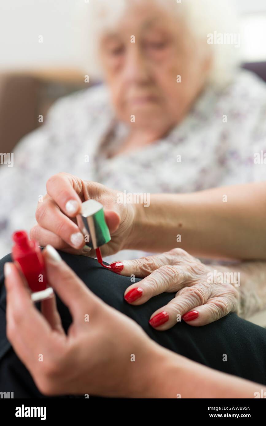 EHPAD - Eine Pflegeassistentin, die nagellack auf die Hände eines älteren und gehörlosen Arztes aufträgt. Stockfoto