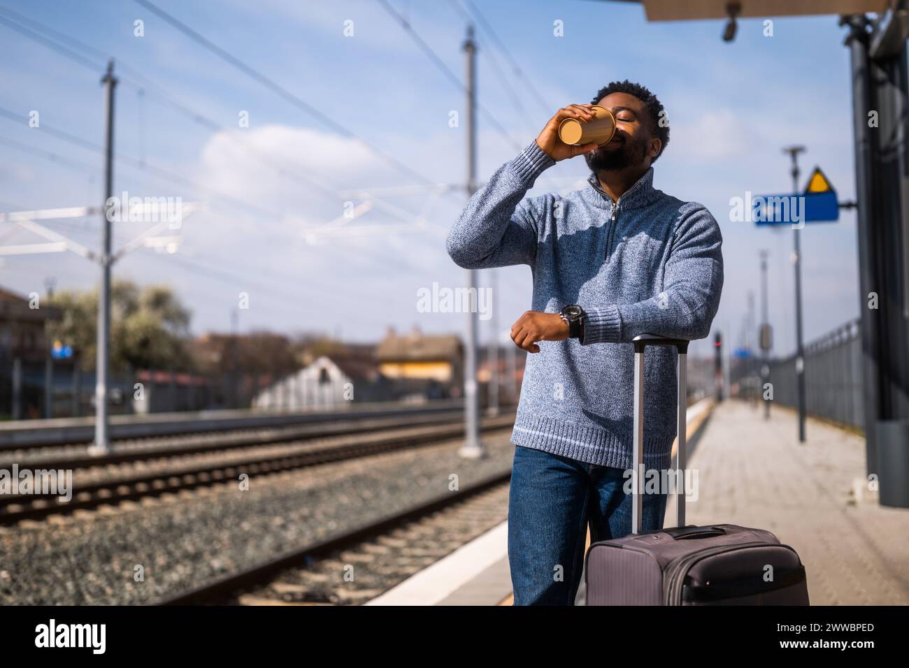 Ein glücklicher Mann mit Koffer trinkt gern Kaffee, während er auf dem Bahnhof steht. Stockfoto