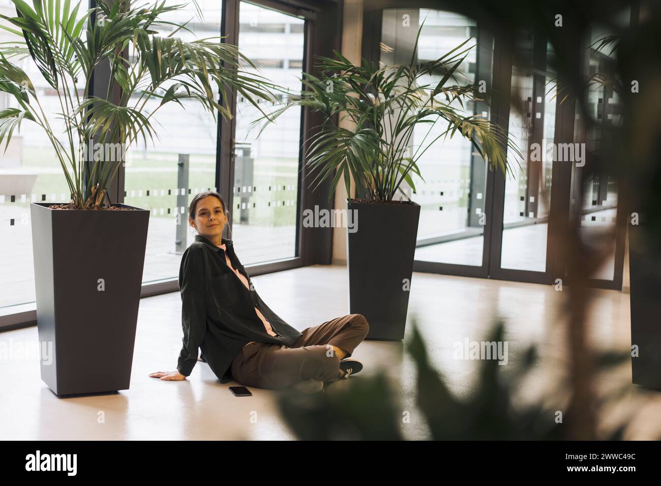 Lächelnde Geschäftsfrau, die neben Pflanzen auf dem Boden sitzt Stockfoto