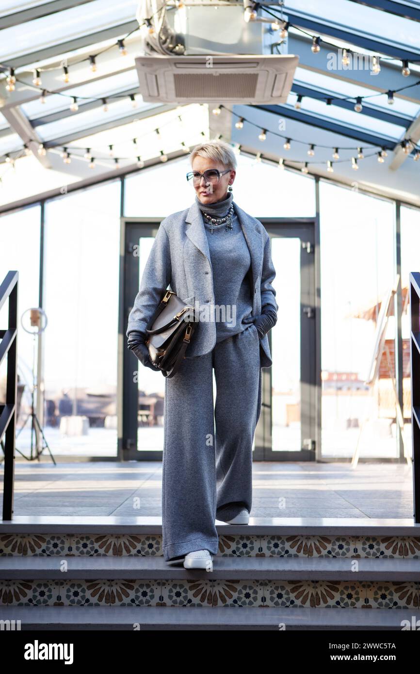 Attraktive und stilvolle reife Frau im grauen Hosenanzug mit Jacke und Rollkragen, mit Handtasche und weißen Schuhen. Der perfekte Look für Succ Stockfoto