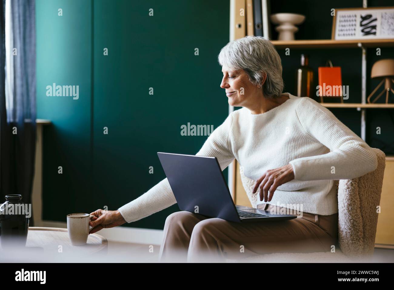 Freiberufler sitzt mit Laptop und nimmt im Home Office Kaffeetasse ab Stockfoto
