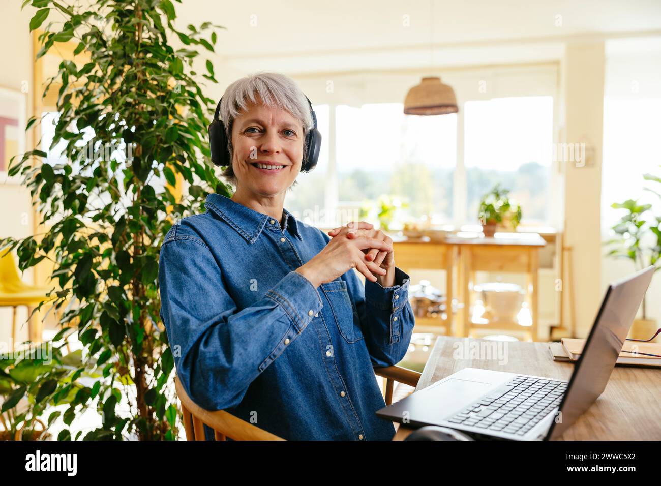 Glücklicher Freelancer, der kabellose Kopfhörer trägt und im Home Office in der Nähe eines Laptops sitzt Stockfoto