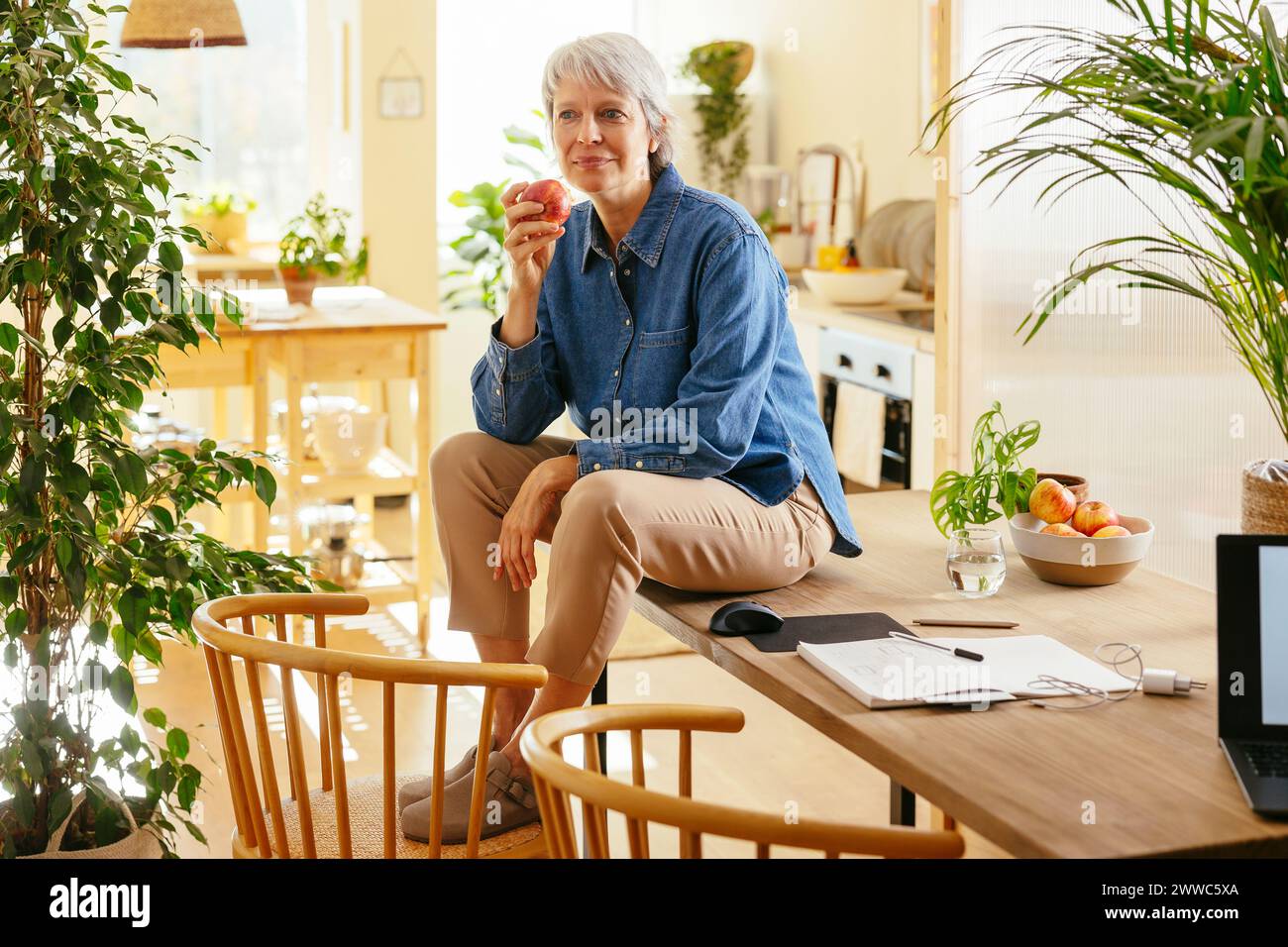 Reifer Freiberufler sitzt auf dem Tisch und hält Apfel im Home Office Stockfoto
