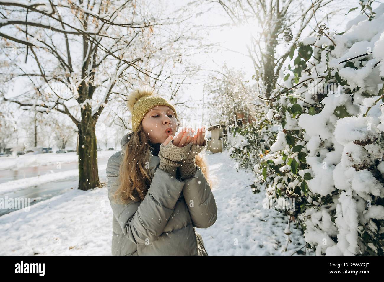 Verspielte Frau, die auf Schnee in der Hand in der Nähe von Pflanzen bläst Stockfoto