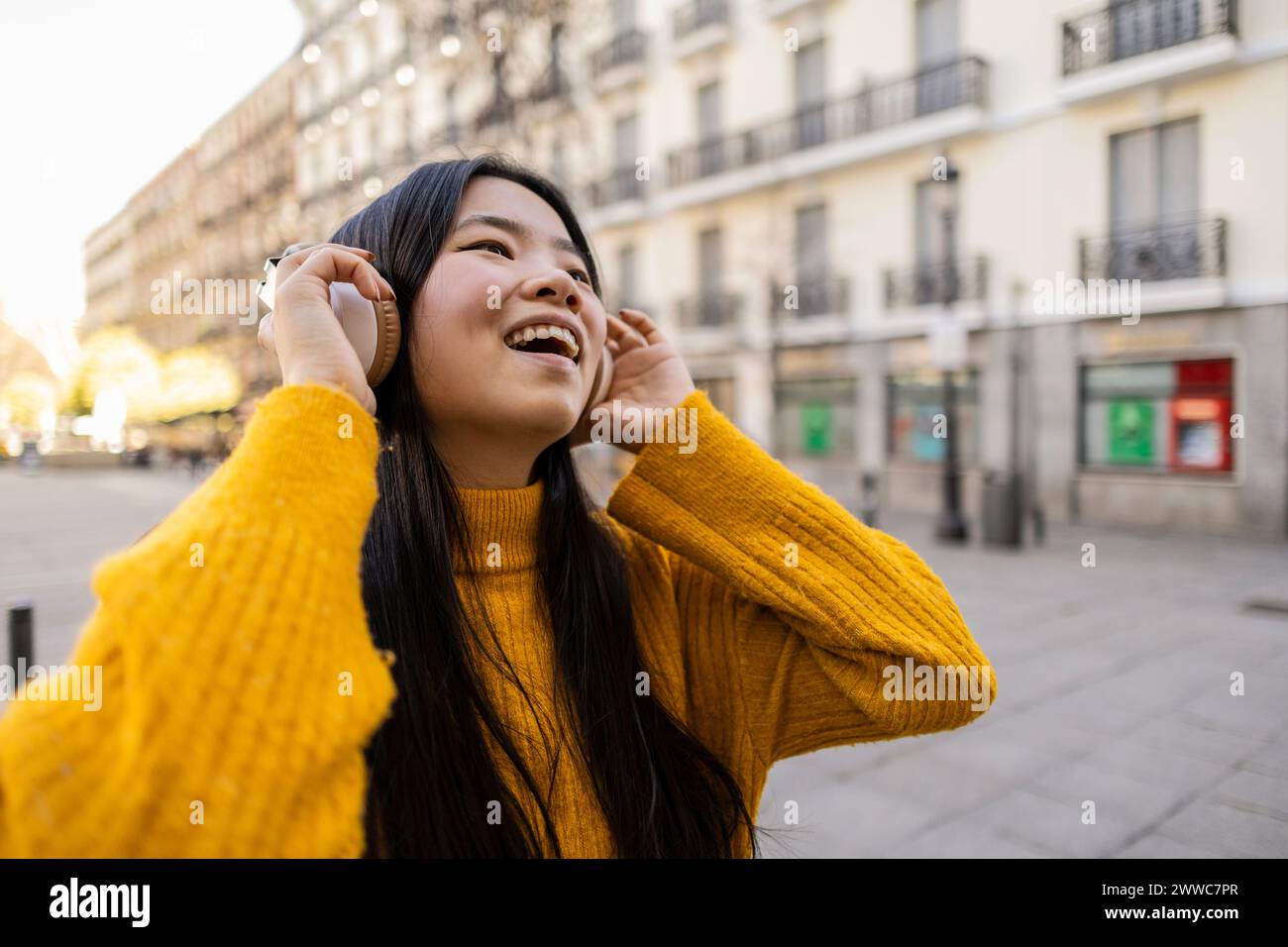 Fröhliche junge Frau, die in der Nähe des Gebäudes kabellose Kopfhörer trägt Stockfoto