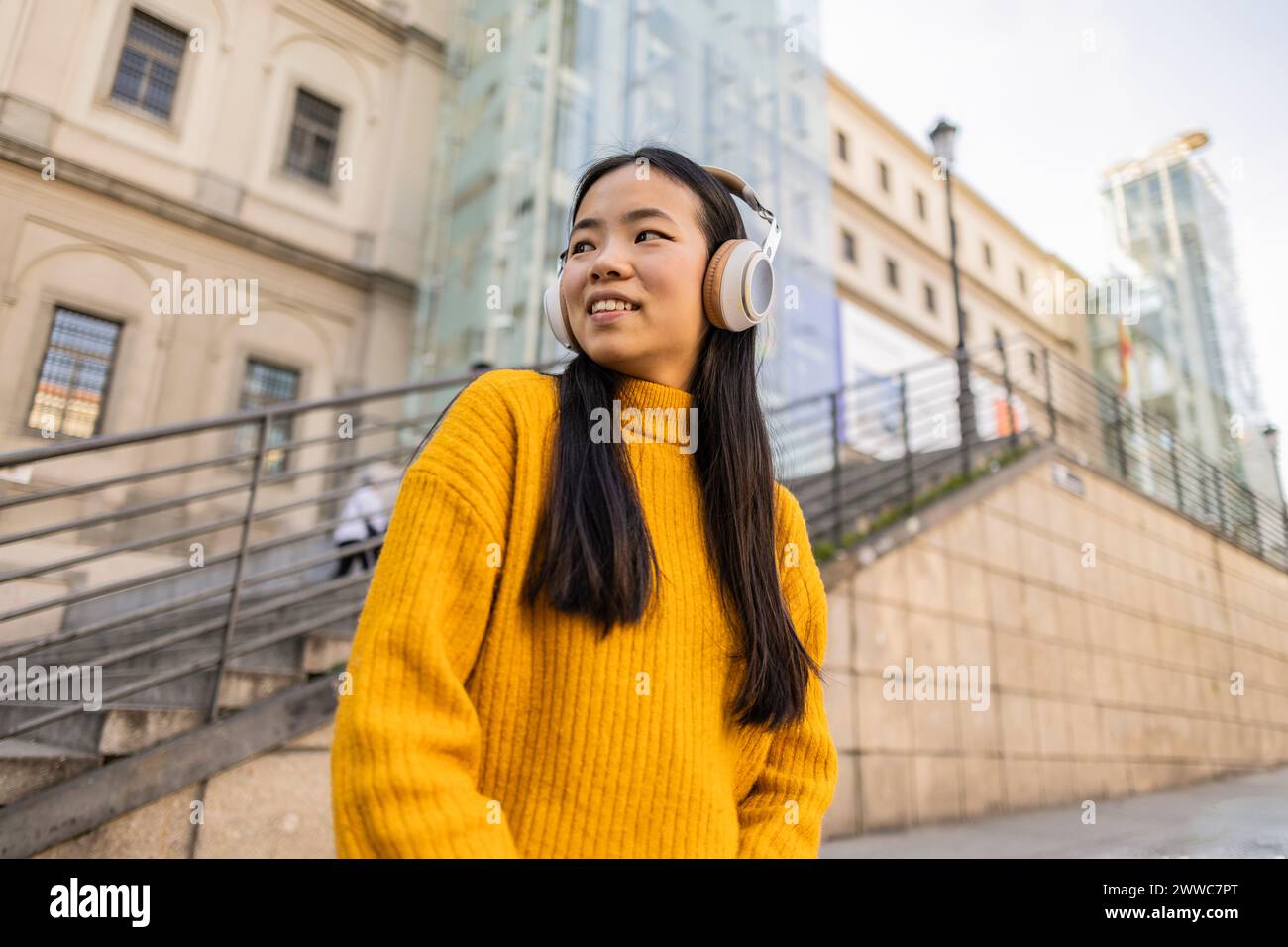 Lächelnde junge Frau, die in der Nähe des Gebäudes kabellose Kopfhörer trägt Stockfoto