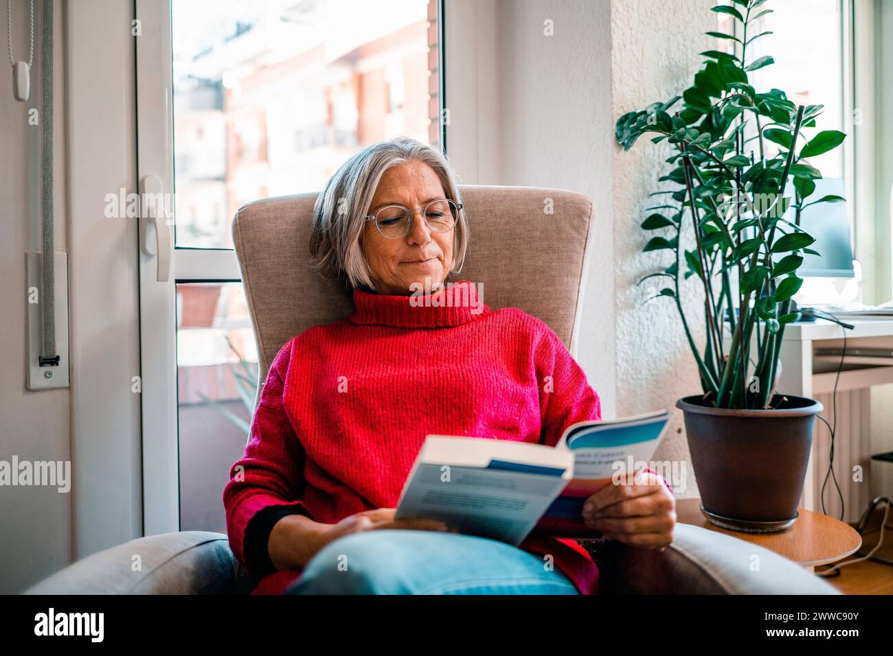 Reife Frau, die zu Hause auf dem Sessel sitzt und Buch liest Stockfoto