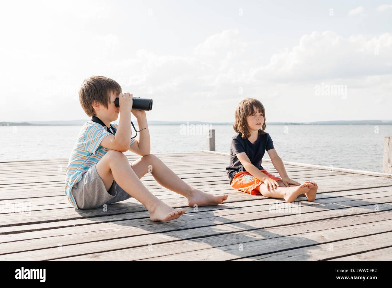 Junge, der durch das Fernglas schaut, sitzt mit Bruder am Pier am See Stockfoto