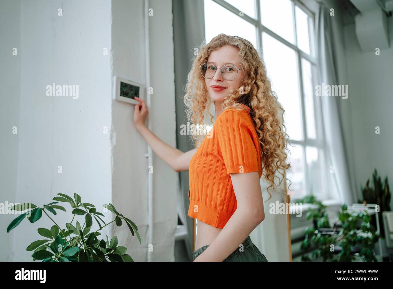 Lächelnde Frau mit blonden Haaren, die zu Hause neben dem Thermostat an der Wand steht Stockfoto