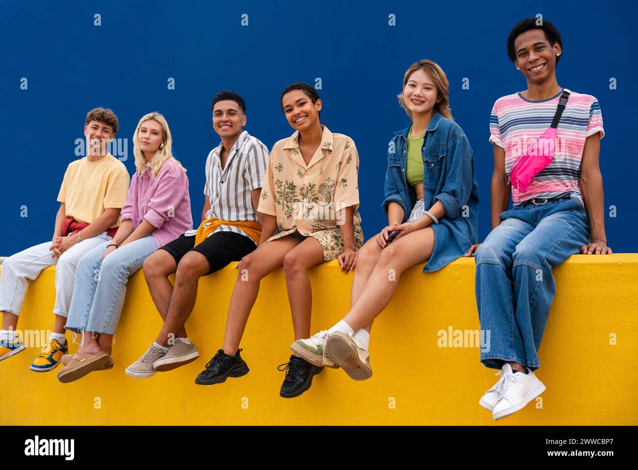 Lächelnde Freunde mit bunten Kleidern sitzen zusammen auf der gelben Wand Stockfoto
