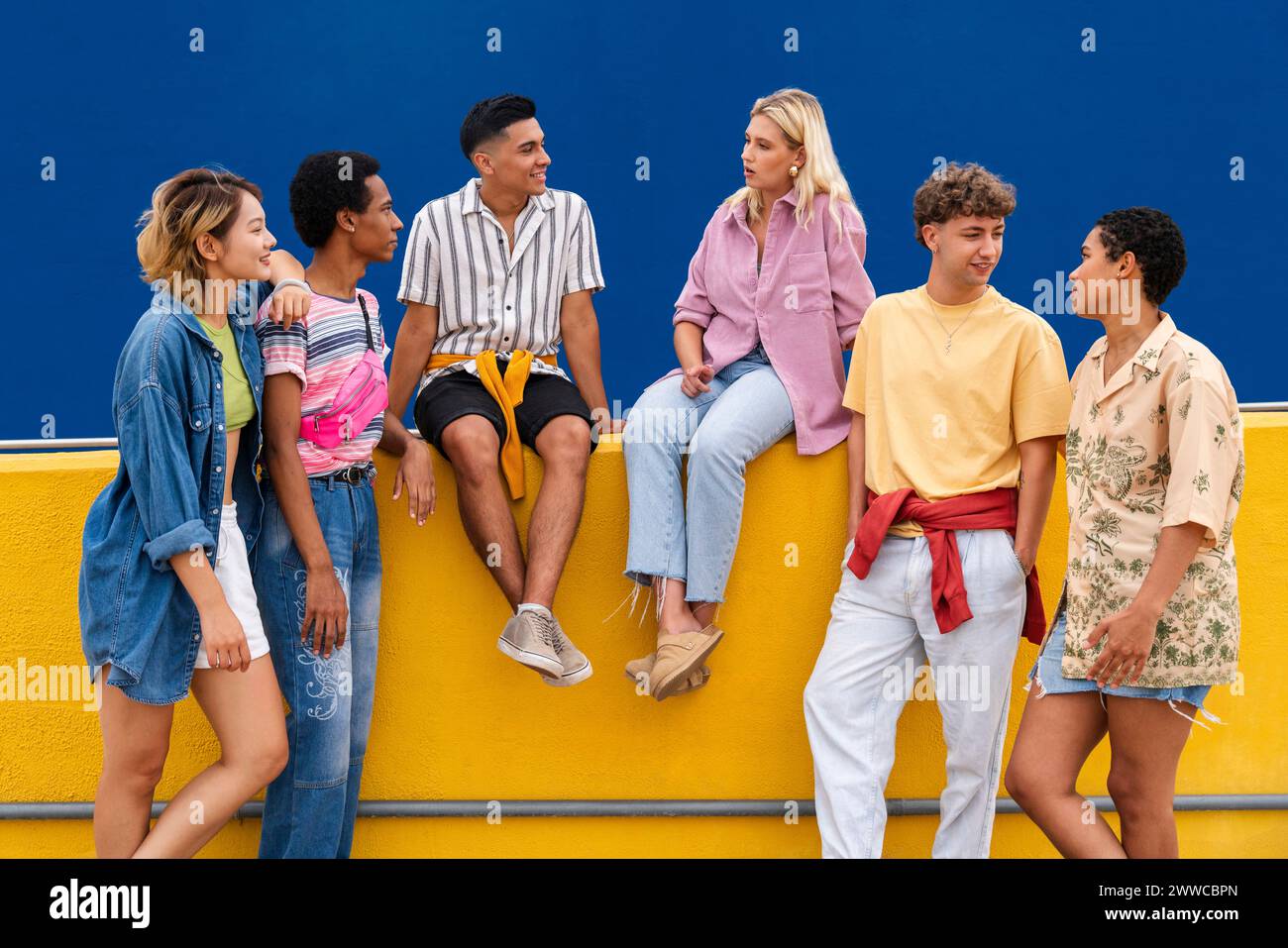 Gruppe von Freunden mit bunten Kleidern, die auf der gelben Wand sitzen und miteinander plaudern Stockfoto