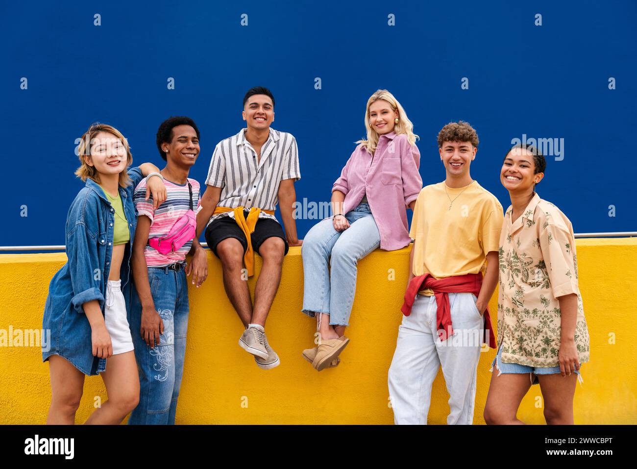 Gruppe von Freunden mit bunten Kleidern, die auf der gelben Wand sitzen und miteinander plaudern Stockfoto